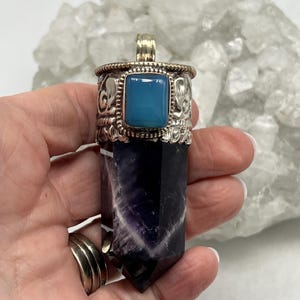May include: A silver pendant with a rectangular, light blue stone and decorative detailing. The pendant is attached to a dark purple and white crystal, with a polished, pointed shape. The pendant is held in a hand.