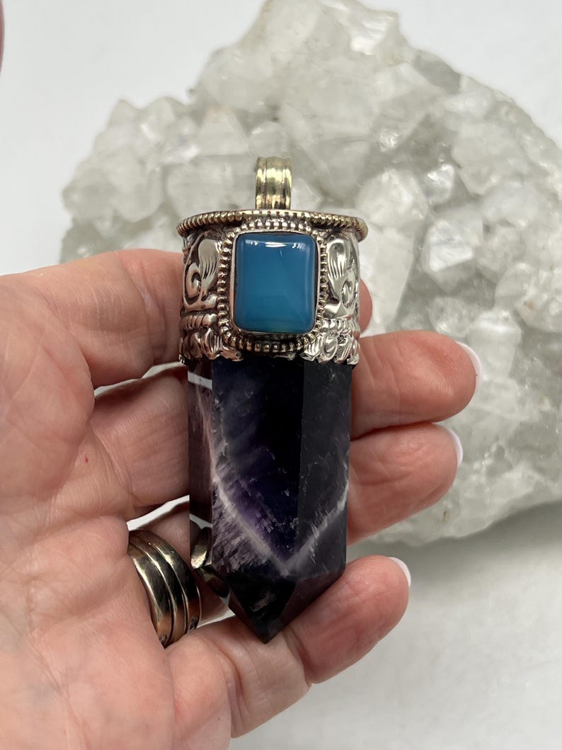 May include: A silver pendant with a rectangular, light blue stone and decorative detailing. The pendant is attached to a dark purple and white crystal, with a polished, pointed shape. The pendant is held in a hand.