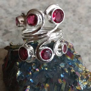 Può includere: Un anello argentato con diverse pietre preziose rosse. L'anello presenta un design a spirale con sei pietre rotonde di un rosso brillante. È esposto su una formazione di cristallo iridescente e colorata, creando un contrasto sorprendente.