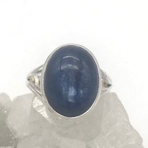 May include: A silver ring featuring a large, oval-shaped, dark blue gemstone. The gemstone is set in a silver band with a split shank design. The ring is displayed on a white, crystalline surface.