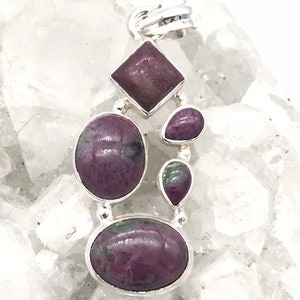 May include: A silver pendant featuring a collection of ruby gemstones in various shapes. The pendant includes a square, oval, and teardrop-shaped stones, all in shades of deep red and purple. The gemstones are set in a silver metal frame.