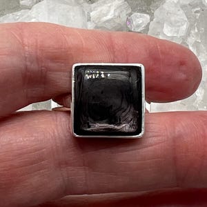 Peut inclure: Une bague en argent avec une pierre carrée noire sertie dans une bélière en argent. La pierre présente un motif tourbillonnant.