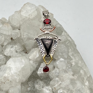 Puede incluir: Un colgante de plata con una piedra triangular negra, rodeada de un marco de plata con detalles dorados. El colgante tiene tres piedras preciosas rojas, una en la parte superior y dos en la parte inferior.