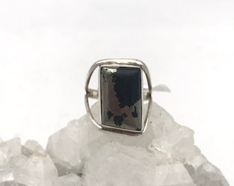 Puede incluir: Un anillo de plata con una piedra de pirita rectangular. La pirita tiene un brillo metálico oscuro con inclusiones más claras y reflectantes. La banda del anillo tiene un diseño simple y abierto, que resalta la forma y el color de la piedra. El anillo está sobre una superficie cristalina blanca.