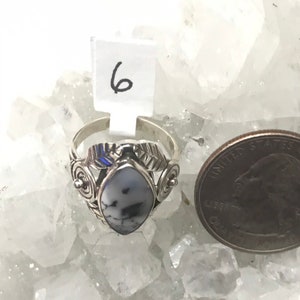 Pu&ograve; includere: Un anello d'argento con una pietra bianca e nera incastonata in un design a foglia. L'anello &egrave; su un letto di cristalli bianchi. Il numero 6 &egrave; visibile su un'etichetta bianca.