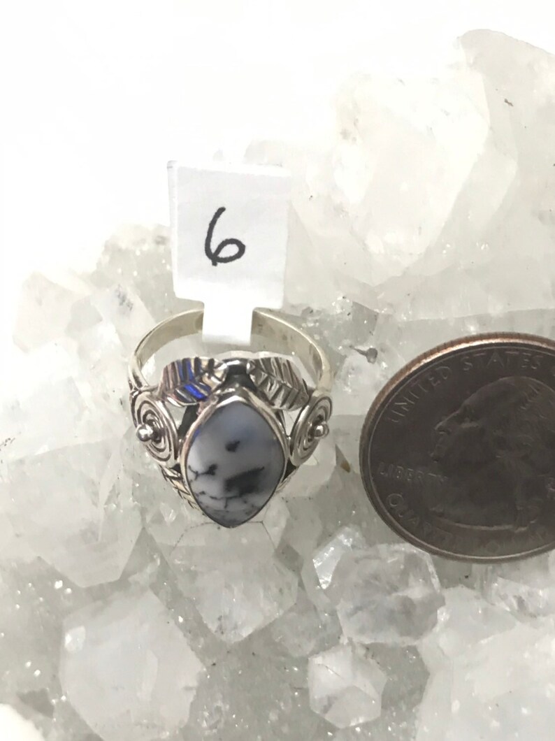 Pu&ograve; includere: Un anello d'argento con una pietra bianca e nera incastonata in un design a foglia. L'anello &egrave; su un letto di cristalli bianchi. Il numero 6 &egrave; visibile su un'etichetta bianca.