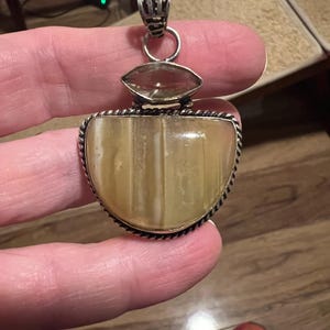 May include: A silver pendant with a large, light-colored, semi-oval stone, and a smaller, clear, oval stone above it. The pendant has a decorative silver bail and a rope-like border around the main stone. The pendant is held by a person.