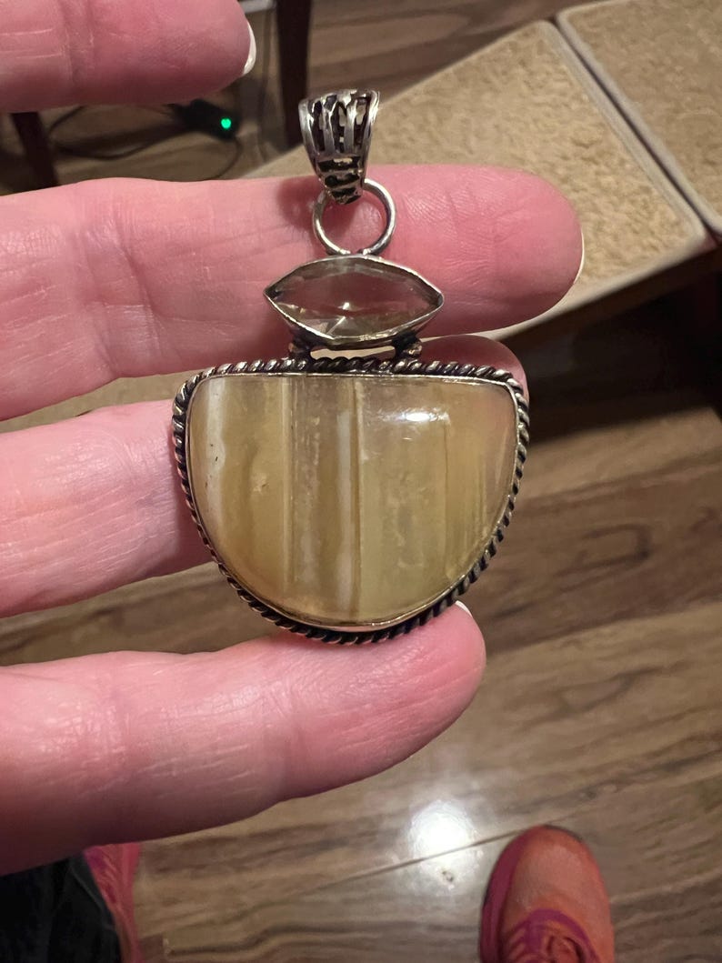 May include: A silver pendant with a large, light-colored, semi-oval stone, and a smaller, clear, oval stone above it. The pendant has a decorative silver bail and a rope-like border around the main stone. The pendant is held by a person.