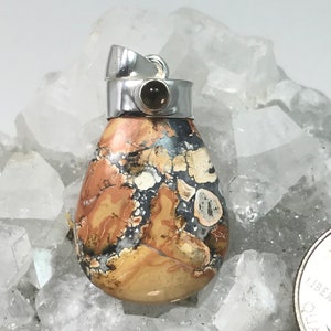 May include: A teardrop-shaped pendant with a silver bail. The pendant features a patterned stone with shades of tan, brown, black, and white. A small, round, dark stone is set in the silver bail. The pendant is displayed on a bed of clear crystals.