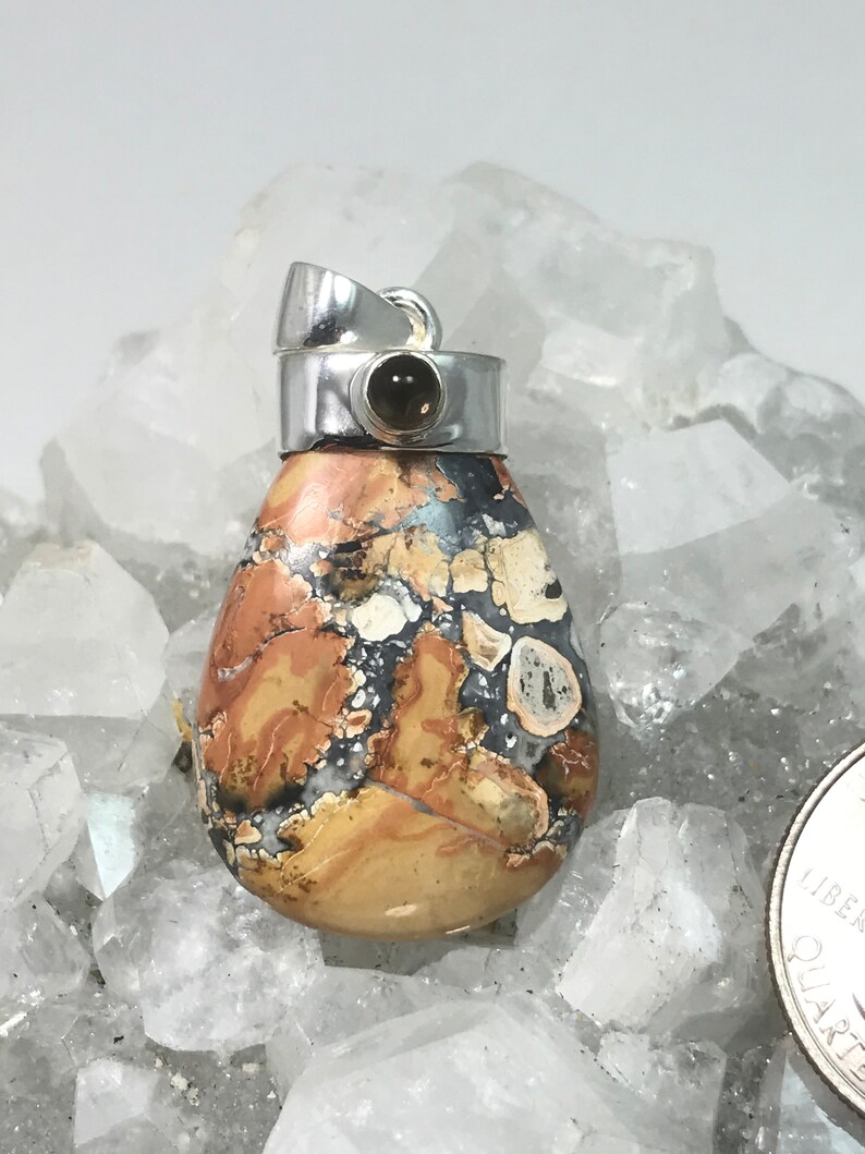 May include: A teardrop-shaped pendant with a silver bail. The pendant features a patterned stone with shades of tan, brown, black, and white. A small, round, dark stone is set in the silver bail. The pendant is displayed on a bed of clear crystals.