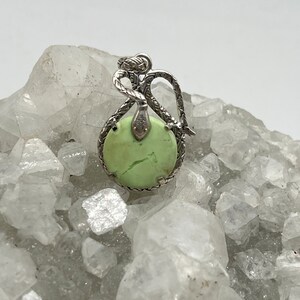 May include: A silver pendant with a green gemstone set in a snake design. The pendant is sitting on a bed of white crystals.