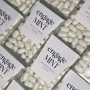 Puede incluir: Cajas de plástico transparente llenas de mentas blancas. Cada caja tiene una etiqueta blanca con las palabras "engage MINT" en negro, junto con los nombres JALAIN & DEJA y la fecha 4.26.25. Las cajas están muy juntas.