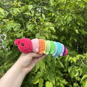May include: A handmade, colourful crochet caterpillar toy. The caterpillar has a red head, followed by segments in pink, orange, yellow, green, light blue, and purple. The toy is held against a backdrop of green foliage.