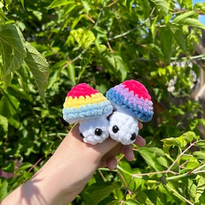 May include: Two crocheted mushroom plushies with rainbow coloured tops and white bodies. The mushrooms have black eyes and are being held in a hand.