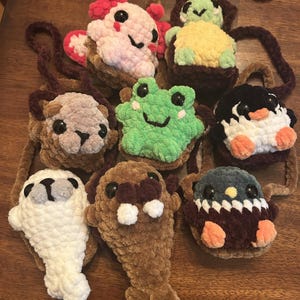Pode incluir: Uma coleção de bolsas de animais de crochê feitas à mão em várias cores. As bolsas apresentam desenhos de axolotl rosa, sapo verde, pinguim, foca, morsa e outras criaturas. Cada bolsa tem uma alça marrom.