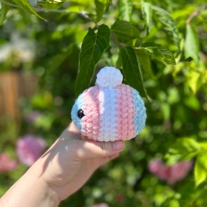 May include: A small, round, crocheted plush toy featuring horizontal stripes in light blue, pink, and white. The plush has a small white pom-pom on top and a single visible black plastic eye on the side, crafted from soft, textured yarn.