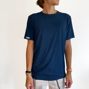 Op de afbeelding: Een man draagt een donkerblauw T-shirt met een klein wit label op de linkermouw. Hij draagt een witte broek en een zilveren ketting.