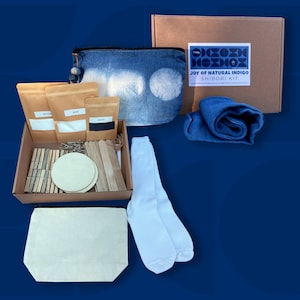 Puede incluir: Un kit Shibori para teñido índigo natural. Incluye una bolsa estilo denim, calcetines blancos, tela azul enrollada, círculos de madera, pinzas y sobres de papel marrón. El kit se presenta en una caja de cartón con la etiqueta "Joy of Natural Indigo Shibori Kit".