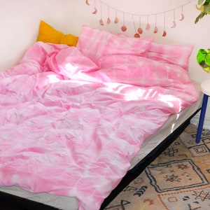 Puede incluir: Ropa de cama de color rosa con teñido anudado con una almohada amarilla en una cama con una pared blanca y una alfombra en el suelo.