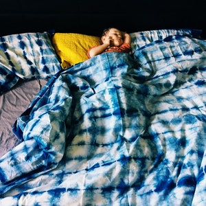 Puede incluir: Un niño duerme en una cama con un edredón y fundas de almohada a juego de color azul y blanco con estampado tie-dye. Se puede ver una almohada amarilla debajo del edredón.
