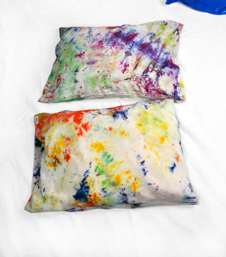 Puede incluir: Dos fundas de almohada de te&ntilde;ido anudado con fondo blanco y patrones de salpicaduras de colores. La funda de almohada de arriba tiene un patr&oacute;n de salpicaduras de morado, rosa, verde y azul. La funda de almohada de abajo tiene un patr&oacute;n de salpicaduras de rojo, naranja, amarillo, verde y azul.