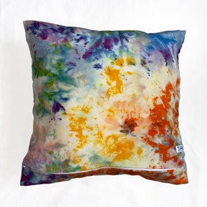 Puede incluir: Una almohada de tie-dye colorida con una cremallera blanca. La almohada tiene tonos de azul, verde, amarillo, naranja y morado.