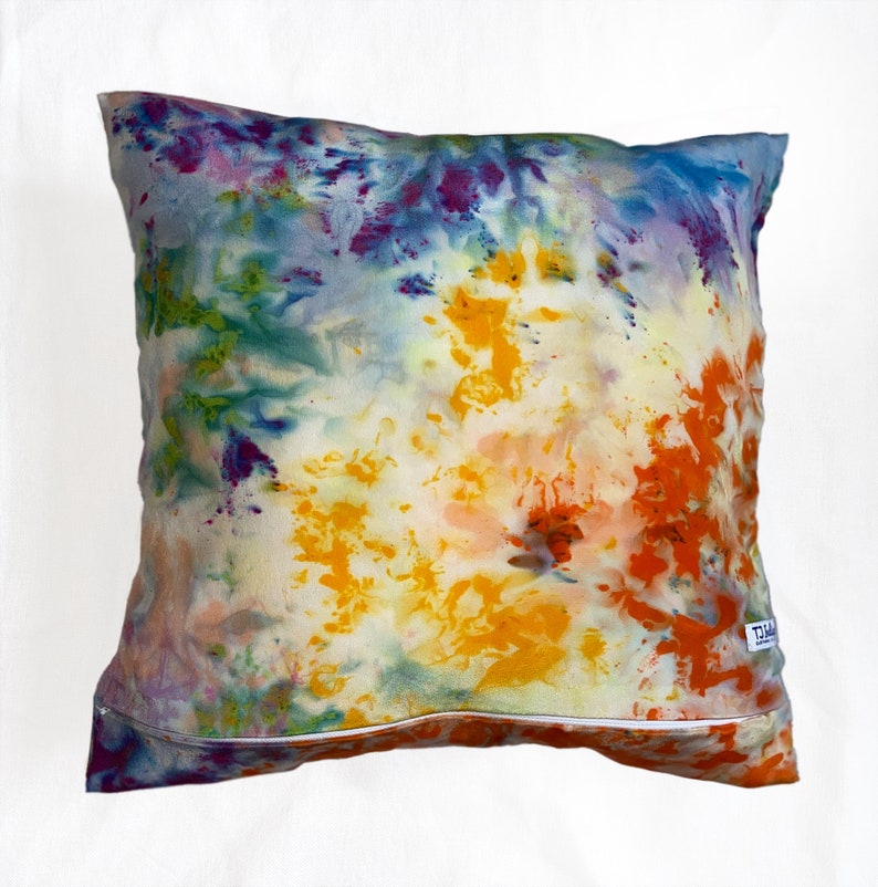 Puede incluir: Una almohada de tie-dye colorida con una cremallera blanca. La almohada tiene tonos de azul, verde, amarillo, naranja y morado.