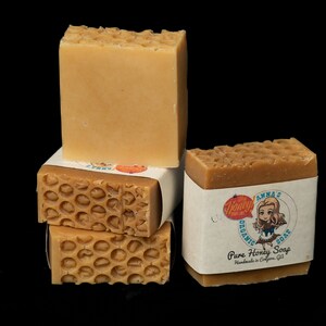 May include: A stack of four honey soap bars. The top bar is plain, while the bottom three bars have a honeycomb pattern. The bottom bar has a label that reads "Anna's Organic Soap, Pure Honey Soap, Handmade in California, USA."
