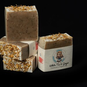 May include: A stack of four bars of handmade soap with a white tea and ginger scent. The soap is wrapped in brown paper with a cartoon illustration of a girl and the text "Anna's Organic Soap White Tea & Ginger Handmade in Oregon, USA".