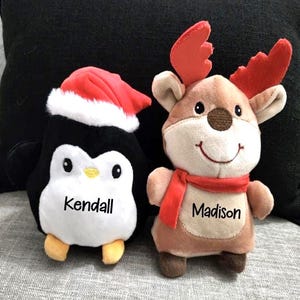 May include: Two plush Christmas toys: a penguin wearing a Santa hat and a reindeer with red antlers and a scarf. The penguin is black and white with the name "Kendall" on its chest. The reindeer has the name "Madison" on its chest.