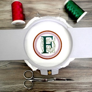 May include: A white embroidery hoop with a green and brown embroidered letter F surrounded by a red and brown circular design. The hoop is on a wooden surface with two spools of thread, one red and one green.