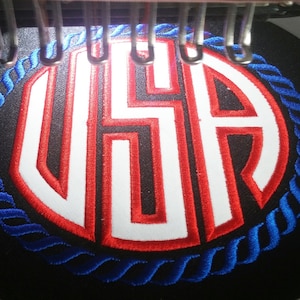 May include: A close-up of a red, white, and blue embroidered patch with the letters "USA" in a circle. The circle is surrounded by a blue rope design.