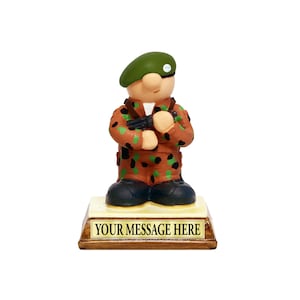 May include: A small figurine of a cartoon soldier wearing a green beret and camouflage uniform. The soldier is holding a gun and is standing on a wooden base with the text "YOUR MESSAGE HERE".