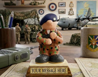 May include: A figurine of a soldier wearing a blue beret and camouflage uniform, standing on a rectangular base with a plaque that reads YOUR MESSAGE HERE. The soldier holds a small black object in their hands.
