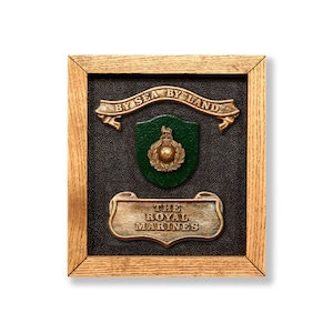 May include: A framed Royal Marines plaque with a dark background and gold and green detailing. The top banner reads "BY SEA BY LAND". The bottom banner reads "THE ROYAL MARINES". The frame is made of light brown wood. A classic military emblem.