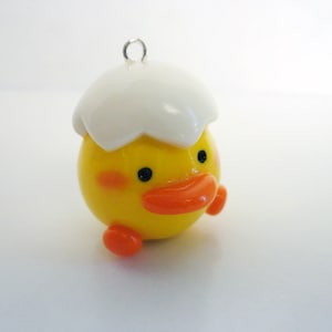 Puede incluir: Un amuleto de pollito amarillo con una parte superior de cáscara de huevo blanca y un pico naranja. El pollito tiene ojos negros y mejillas rosadas.