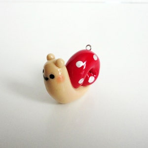 May include: A small, beige snail charm with a red and white polka dot shell. The snail has a cute face with black eyes and a small smile.