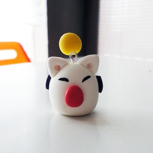 May include: White and red clay charm with a yellow ball on top. The charm has a small, black, circular detail on each side of the head.