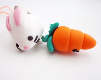 May include: A white bunny charm with pink ears and black eyes, attached to a carrot charm with green leaves and orange body.