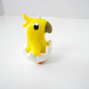 May include: A yellow chick figurine with a brown beak and black eyes, emerging from a white eggshell.