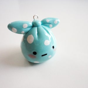 May include: A small blue and white polka dot clay charm with a cute face and a bow. The charm is shaped like a round ball with a small loop at the top for hanging.