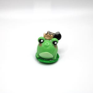 Op de afbeelding: Een groene kikkerfiguur met een gouden kroon en zwart accessoire. De kikker heeft grote ogen en zit op een groen lelieblad. Een grillig decoratief item.
