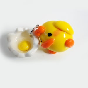 May include: A yellow chick charm with orange accents, holding a white egg with a yellow yolk. The chick and egg are made of polymer clay.
