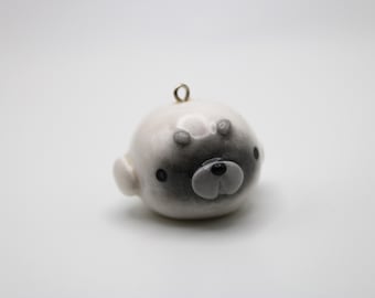 May include: A round, white ceramic pendant shaped like a seal head with a glossy finish. The piece features a dark grey gradient around the eyes and nose, with small black dots for eyes and a small metal loop at the top for hanging as a charm.