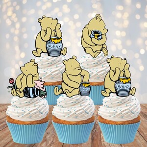 May include: Five cupcakes in light blue paper liners topped with white frosting and blue sprinkles. Each cupcake features a Winnie the Pooh character cutout, with some bears holding honey pots labeled HUNNY or sitting with a small pink piglet figure.