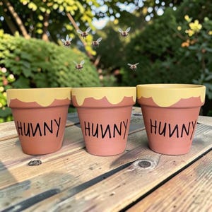 May include: Three terracotta flower pots painted with a yellow drip pattern at the rim and the word HUNNY written in black lettering on the front. The pots are arranged in a row, and several small bee illustrations are floating above them.