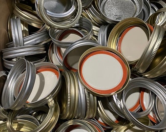 May include: A collection of silver and gold-colored jar lids and rings. The lids have a white interior with a red rubber seal. The rings are made of metal and are designed to screw onto the jars. The lids and rings are in a cardboard box.