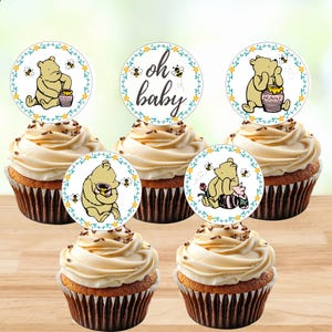 May include: Five cupcakes topped with circular paper decorations featuring Winnie the Pooh illustrations, honey pots, bees, and floral borders. One topper displays the text oh baby in black script, while others show the character in various poses.