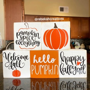 May include: A collection of five wooden blocks with fall-themed designs. The blocks feature phrases like "Pumpkin Spice Everything" and "Welcome Fall," along with pumpkin illustrations in orange and black. The blocks are arranged on a reflective surface.