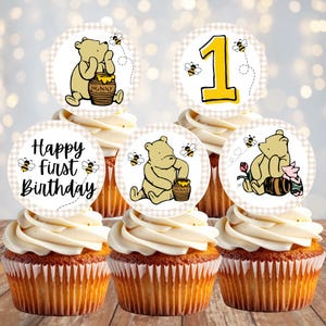 May include: Five cupcakes topped with circular paper toppers featuring Winnie the Pooh, Piglet, honey pots, and bees. The designs include the number 1 and the text Happy first Birthday, all set against a light beige checkered border pattern.